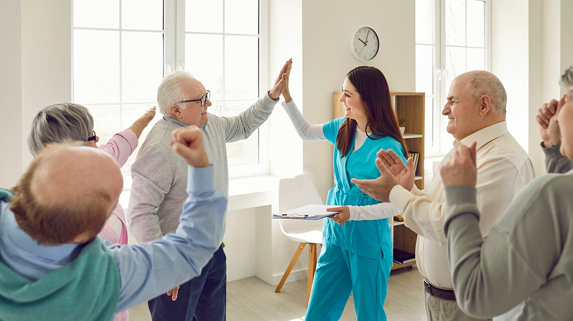 Pflegerin gymnastiziert mit Senior:innen in einem Pflegeheim. Mit einem der Senioren macht sie einen "High Five"-Abschlag, Brain City Berlin 