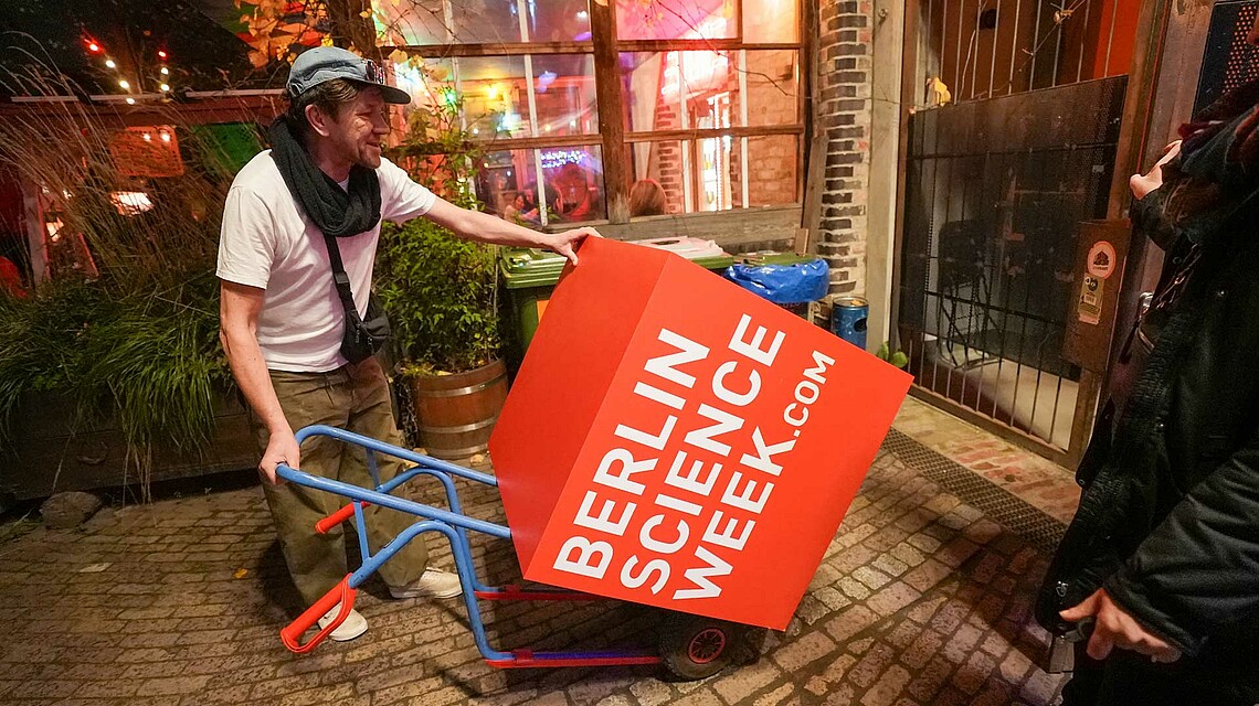 Man with wheelbarrow pushes red Berlin Science Week logo cube, Brain City Berlin