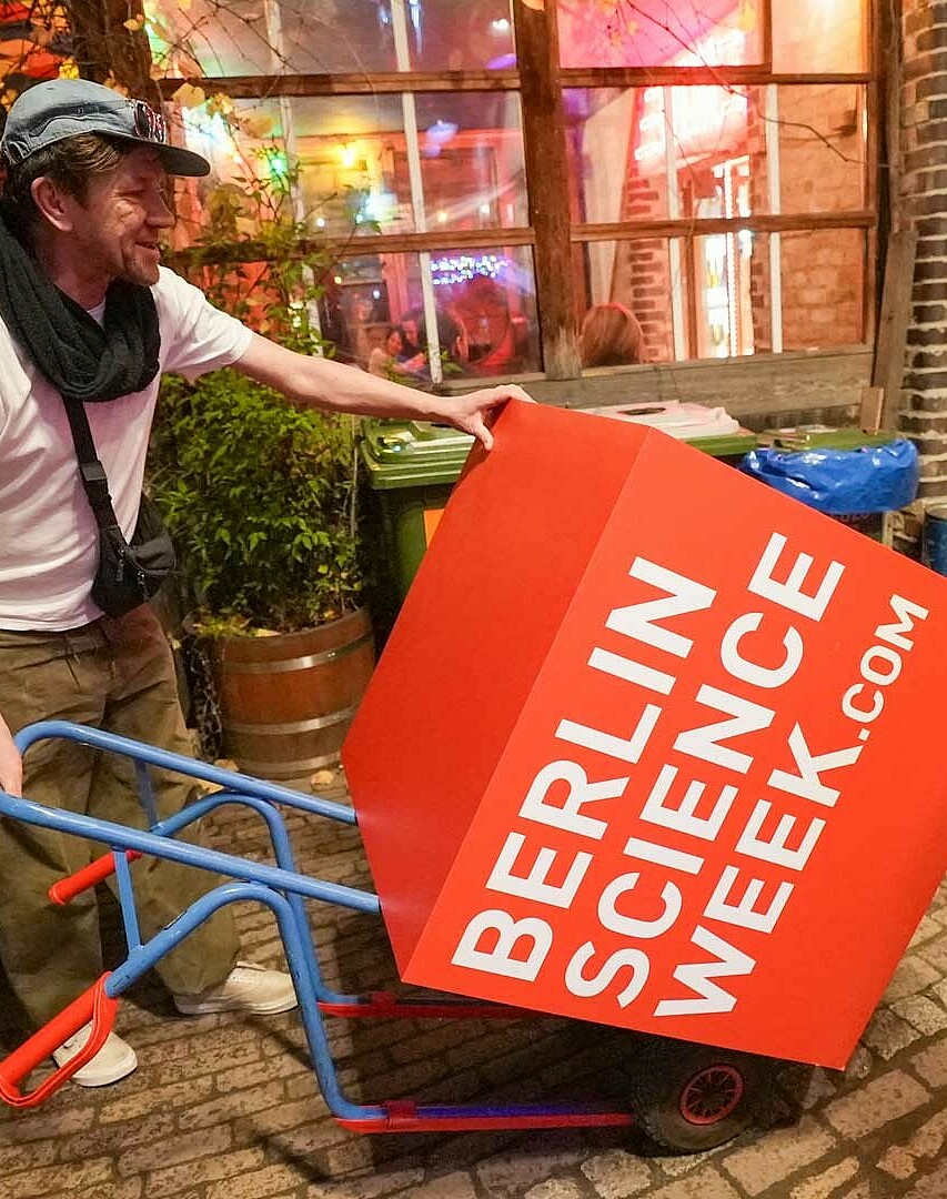 Man with wheelbarrow pushes red Berlin Science Week logo cube, Brain City Berlin